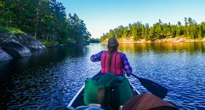 a young person with a long braid sits in the front of a canoe holding a paddle, facing away from the camera. There are evergreen trees lining the shore.